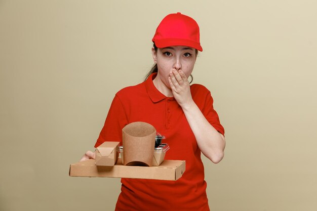赤い帽子と空白の t シャツの制服を着た配達の女性従業員は、茶色の背景の上に立って手で口を覆っている驚きとショックを受けたカメラを見て食品箱を保持しています