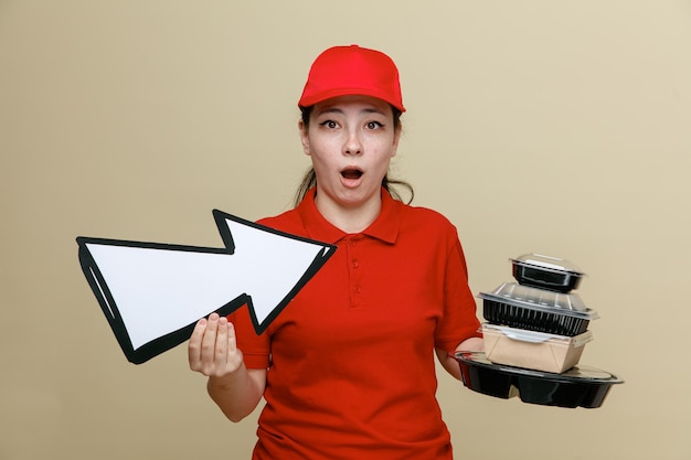 Delivery woman employee in red cap and blank tshirt uniform holding food containers and big arrow looking at camera amazed and surprised standing over brown background