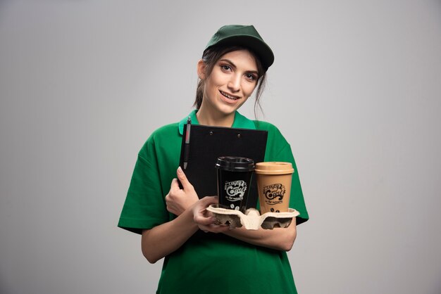 Delivery woman in green uniform holding clipboard and coffee cups. 