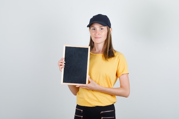 Donna di consegna tenendo la lavagna in t-shirt, pantaloni e berretto e guardando felice