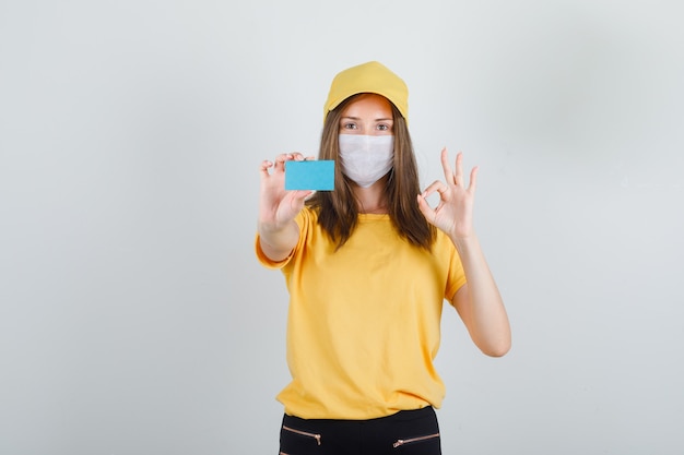 Delivery woman holding blue card with ok sign in t-shirt, pants and cap, mask and looking glad