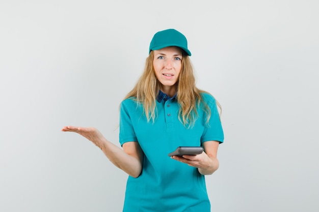 Delivery woman holding calculator with spread palm in t-shirt, cap and looking confused 