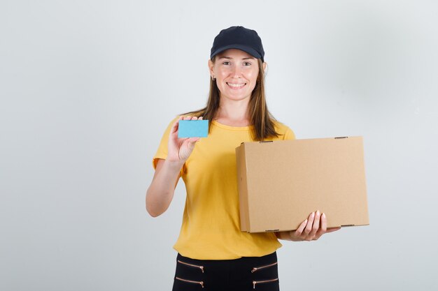 Delivery woman holding cardboard box and blue card in t-shirt, pants and cap and looking glad