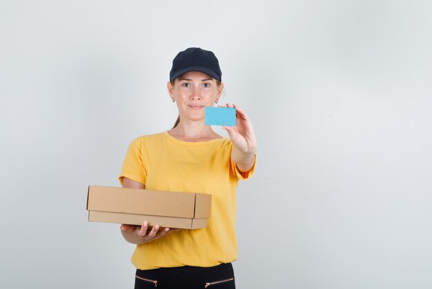 Delivery woman holding cardboard box and blue card in t-shirt, pants and cap