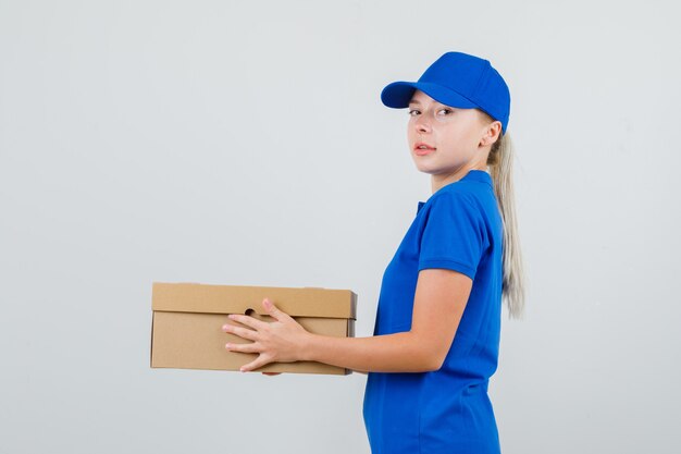 Delivery woman holding cardboard box in blue t-shirt and cap and looking confident .