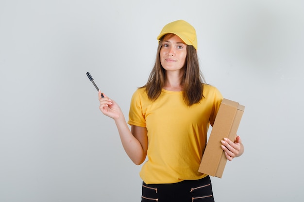 Donna di consegna che tiene scatola di cartone e penna in t-shirt, pantaloni e berretto e sembra felice