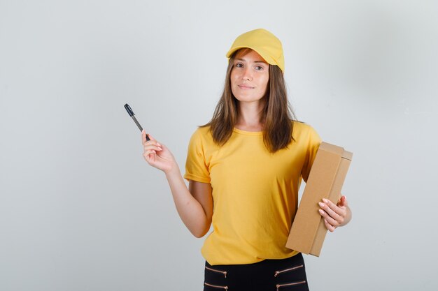 Доставщица держит картонную коробку и ручку в футболке, штанах и кепке и выглядит довольной