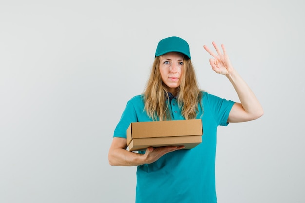 Delivery woman holding cardboard box and showing ok sign in t-shirt, cap 