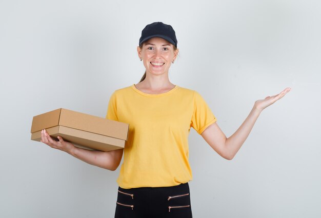 Доставщик женщина держит картонную коробку и улыбается в футболке, штанах и кепке