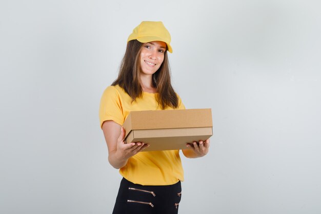 Доставщица держит картонную коробку и улыбается в желтой футболке, штанах и кепке