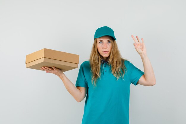 Женщина доставки, держащая картонную коробку с одобренным жестом в футболке, крышке и уверенно выглядящая.