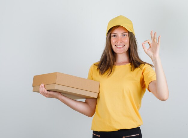 Delivery woman holding cardboard box with ok sign in yellow t-shirt, pants and cap and looking glad