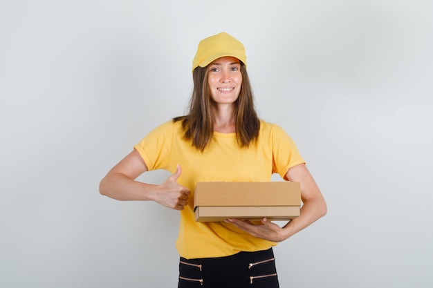 Donna di consegna tenendo la scatola di cartone con il pollice in su in t-shirt, pantaloni e berretto e guardando allegro