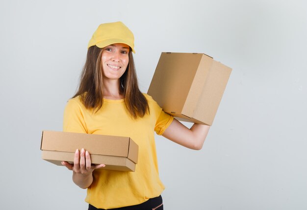 Доставщик женщина держит картонные коробки и улыбается в желтой футболке, штанах и кепке