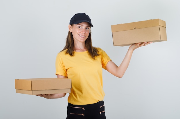 Доставщица держит картонные коробки в футболке, штанах и кепке и выглядит веселой