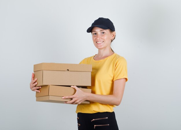 Delivery woman holding cardboard boxes in t-shirt, pants and cap and looking happy