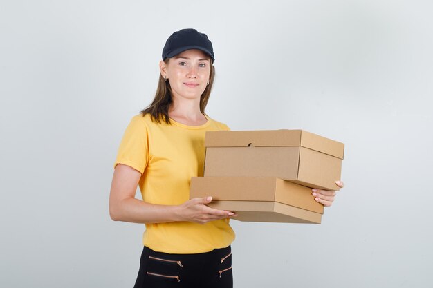Delivery woman holding cardboard boxes in t-shirt, pants and cap and looking jolly