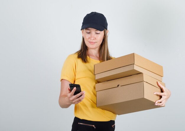 Delivery woman holding cardboard boxes and using smartphone in t-shirt, pants and cap