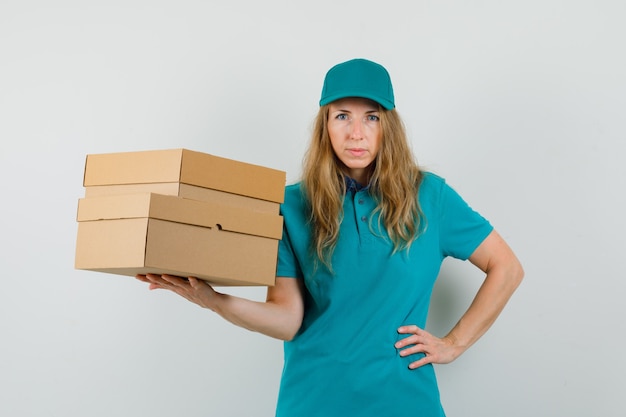 Delivery woman holding cardboard boxes with hand on waist in t-shirt, cap 