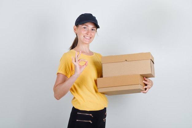 Женщина-доставщик, держащая картонные коробки с одобренным знаком в футболке, штанах, кепке и выглядящей веселой.