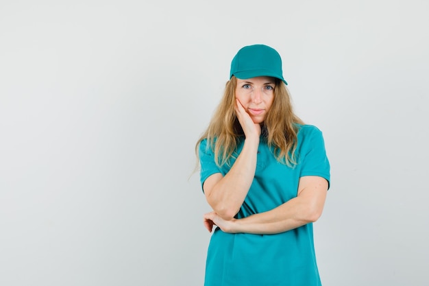 Delivery woman holding hand on cheek in t-shirt, cap and looking sensible. 