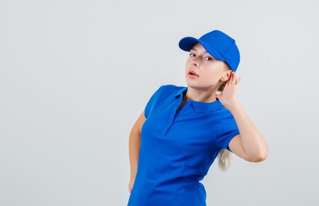 Delivery woman holding hand behind ear in blue t-shirt and cap and looking curious