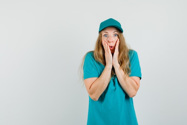 Delivery woman holding hands near mouth in t-shirt, cap and looking shocked 