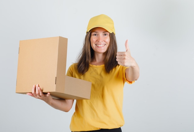 Delivery woman holding opened box with thumb up in yellow t-shirt, pants, cap and looking cheery