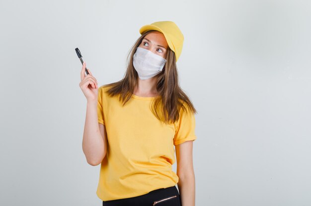 Delivery woman holding pen and looking up in t-shirt, pants, cap, mask and looking careful