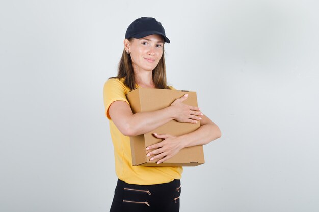 Delivery woman hugging cardboard box in t-shirt, pants, cap and looking glad.