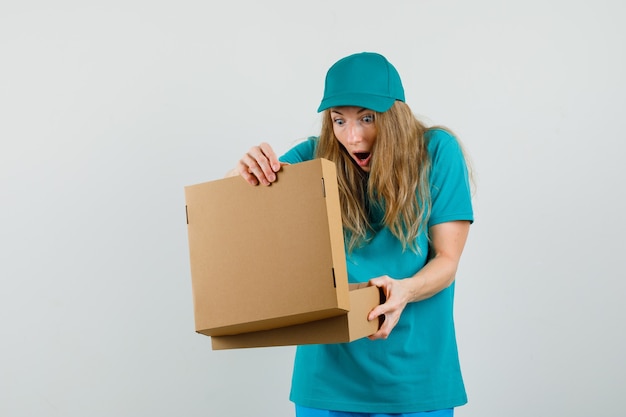 Delivery woman looking into cardboard box in t-shirt, cap and looking wondered. 