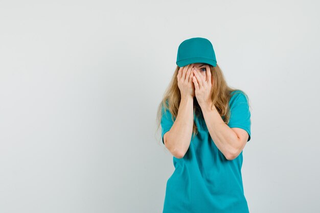 Free photo delivery woman looking through fingers in t-shirt, cap and looking scared.