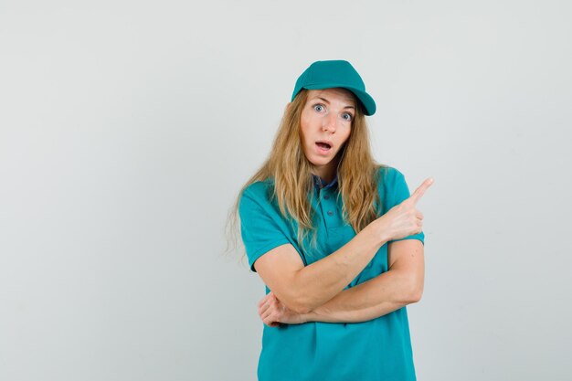 Delivery woman pointing away in t-shirt, cap and looking amazed 