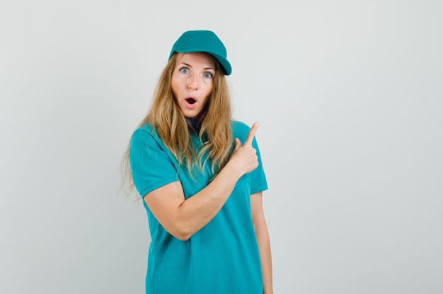 Delivery woman pointing away in t-shirt, cap and looking surprised 