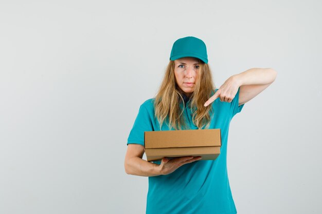 Delivery woman pointing at cardboard box in t-shirt, cap 