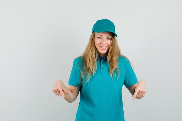 Delivery woman pointing down in t-shirt, cap and looking cheerful. 