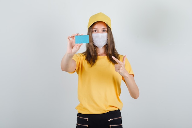 Delivery woman pointing finger at blue card in t-shirt, pants, cap and mask and looking glad