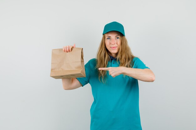 Delivery woman pointing at paper bag in t-shirt, cap and looking cheery. 