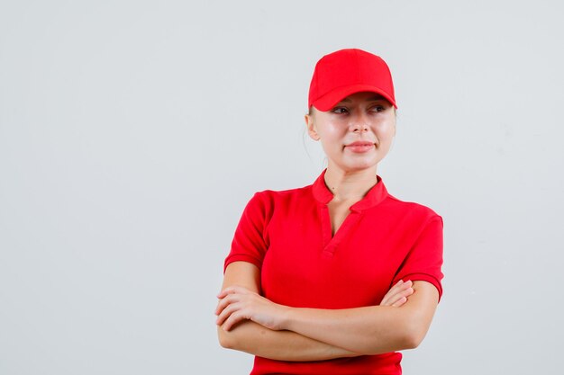 Женщина-доставщик в красной футболке и кепке смотрит в сторону со скрещенными руками и выглядит с надеждой
