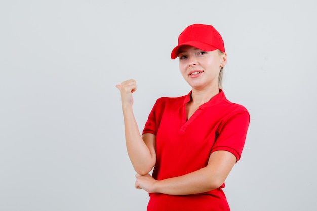 Женщина-доставщик в красной футболке и кепке, указывая большим пальцем назад и выглядит уверенно