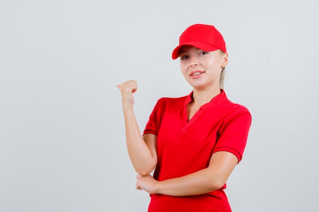 Delivery woman in red t-shirt and cap pointing back with thumb and looking confident