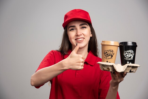 Женщина-доставщик в красной форме держит кофейные чашки и делает большие пальцы руки вверх.