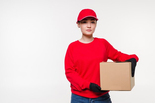 Delivery woman in red uniform isolated on white wall. Courier in medical gloves, cap, red t-shirt working as dealer holding cardboard box to deliver. Receiving package.