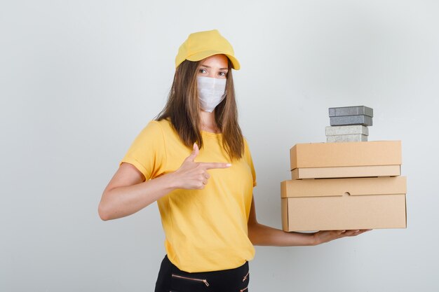 Delivery woman showing boxes with finger in t-shirt, pants, cap and mask and looking cheery