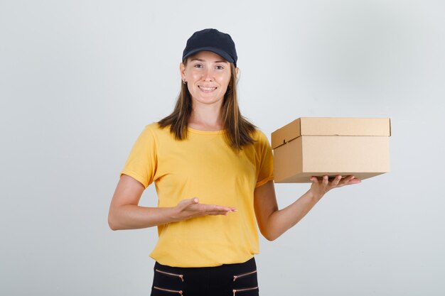 Delivery woman showing cardboard box in t-shirt, pants, cap and looking jolly