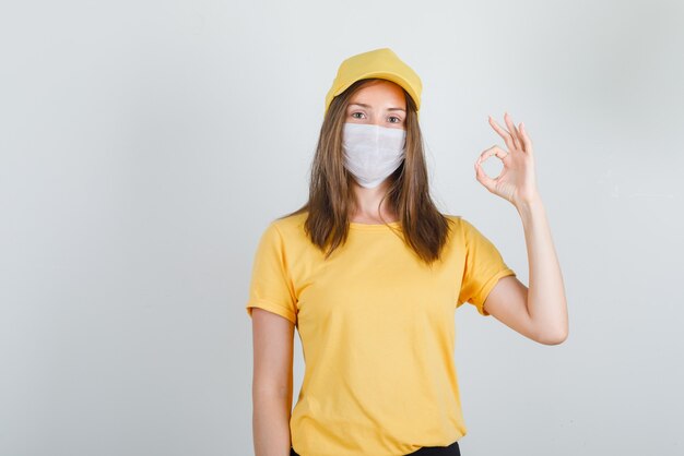 Delivery woman showing ok gesture in t-shirt, pants, cap and mask and looking glad.