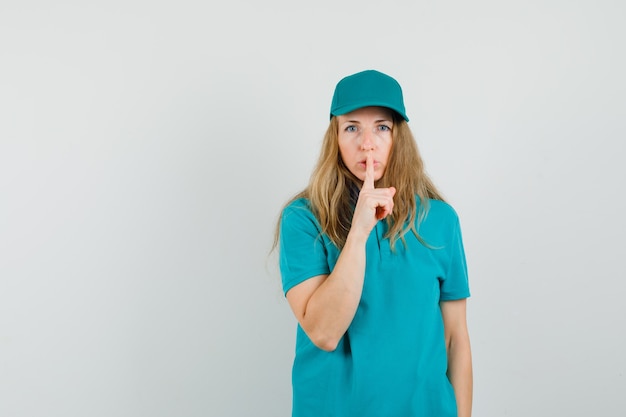 Delivery woman showing silence gesture in t-shirt, cap and looking careful 