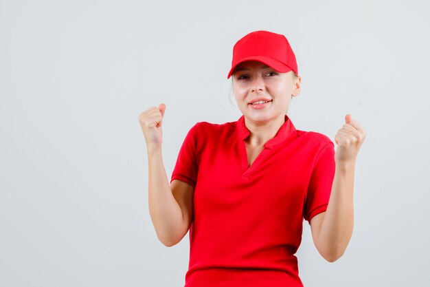 Женщина-доставщик показывает жест победителя в красной футболке и кепке и выглядит счастливой