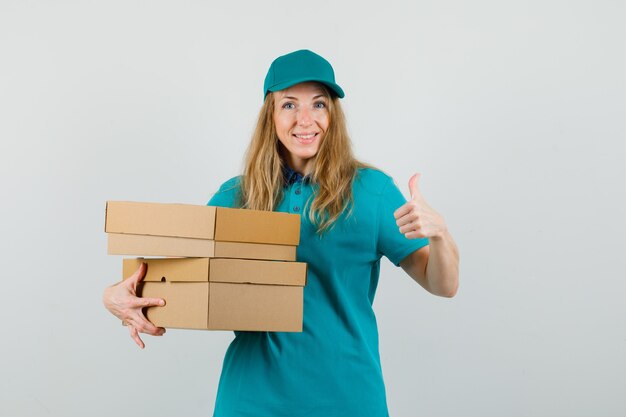 Delivery woman in t-shirt, cap holding cardboard boxes with thumb up and looking glad 