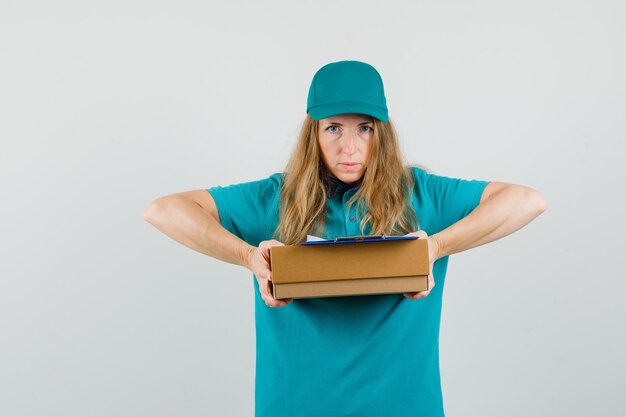 Delivery woman in t-shirt, cap holding clipboard on cardboard box and looking serious 